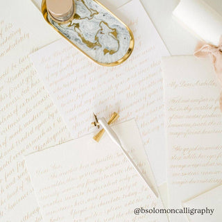 Several sheets of handwritten calligraphy are displayed on a white surface with the Flourish Calligraphy Pen, an ink bottle on a marble tray, and a rolled-up ribbon. The handle @bsolomoncalligraphy is shown at the bottom.