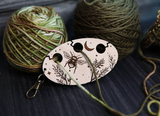 The Yarn Management Tool for Knitters, featuring a bee, moon, and leaf design, is pictured resting between two balls of green yarn on a dark surface, with yarn threaded through one of its holes.