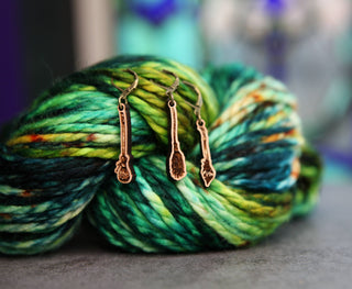 Three Witch’s Broom Stitch Markers hang on a skein of thick, multicolored yarn in green, yellow, blue, and teal—perfect accessories for knitting or crochet projects. The blurred background keeps the focus on these magical markers.