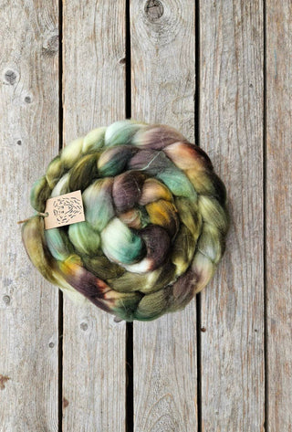 The Koigu Merino Roving Braid, a coiled blend of green, brown, yellow, and gray fiber, rests on weathered wood with visible knots and grain. A small paper tag is tucked into the spinning fiber.