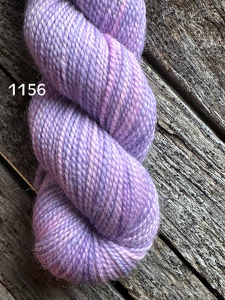 A skein from the KPM and KPPPM Variegated Color Collection is coiled on weathered wood, displaying soft purple hues. The number 1156 appears in white, ideal for hand knitting enthusiasts seeking premium merino yarn.