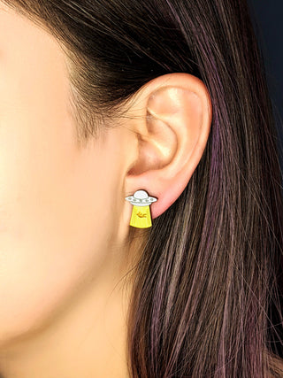 A close-up shows a person with straight brown hair tucked behind their ear, wearing Alien Joyride Earrings—whimsical, handmade UFOs beaming yellow light. The unique earrings are hypoallergenic and stand out in the shot.