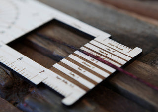 Close-up of a Butterfly "Almost Everything" Ruler measuring yarn thickness, with clear markings and labels visible, resting on a rustic wooden surface.