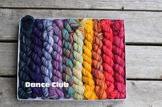 A Koigu Pencil Box in the Dance Club colorway displays merino wool yarn skeins arranged in rainbow order on a wooden surface, with “Dance Club” in bold white letters at the lower left.