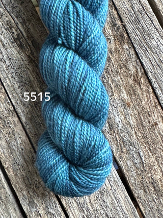 A skein of Koigu KPM & KPPPM Fingering Weight Yarn in hand-dyed blue (label 5515) sits on a rustic wooden surface, ideal for knitting and crochet projects.