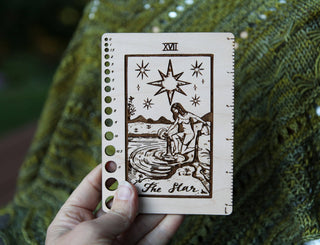 A hand holds the Maple Wood Tarot Card Knitting Needle Gauge & 5 Ruler, featuring The Star design with a woman pouring water, stars above, and ruler holes along the edge, set against textured green fabric.