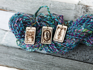 Three engraved Tarot Card Stitch Markers—The World, The Lovers & The Hanged Man—rest on blue, purple, and green sequin yarn beside maple wood knitting progress keepers on a rustic wooden surface.