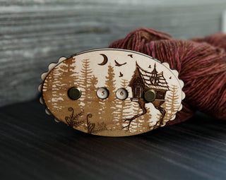 The Baba Yaga Row Counter, an oval wooden tool engraved with a folklore-inspired forest and house design, displays numbered dials reading 5 and 6 as it rests on dark wood beside a skein of reddish yarn.