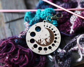 The Crescent Moon Knitting Needle Gauge, crafted from cherry wood and featuring a mountain design, sits atop colorful yarn and pink knitting needles. Its round shape has numbered holes to measure different needle sizes.