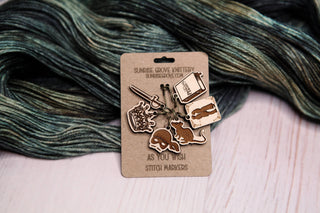 The Princess Bride Knitting Stitch Markers are a set of maple wood stitch markers featuring fantasy-themed designs such as a sword, crown, dinosaur, book, and ball of yarn. Displayed on a branded card against dark green and gray yarn, these charming knitting accessories add a whimsical touch to your craft projects.
