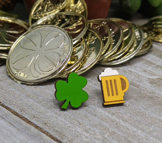A pile of gold coins rests on a wooden surface, with two small earrings in front—one shaped like a green clover and the other like a foaming beer mug. These handmade-in-USA Clover and Beer Earrings add a unique, festive touch.