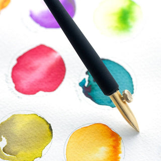 The One-Dip-Nib, a gold-accented black fountain pen with a calligraphy nib, lies on watercolor paper adorned with vibrant red, green, teal, yellow, and orange paint swatches.