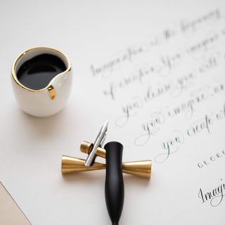 The Flourish Calligraphy Pen and a metal nib rest beside a white cup of black ink on handwritten paper, creating a calm, creative writing atmosphere.