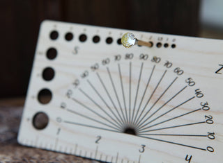 A close-up of the Apothecary "Almost Everything" Ruler—a wooden tool with circular holes, numbered markings, angle lines, and a jeweled pin at the top casting a shadow on its engraved scale.