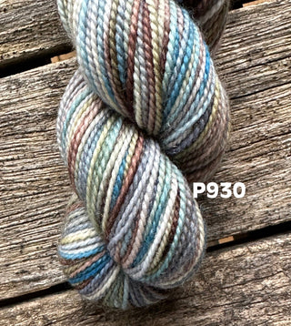 A skein from the KPM and KPPPM Variegated Color Collection in brown, gray, blue, and green tones rests on weathered wood. The code P930 is shown beside this soft merino wool—ideal for hand knitting projects.