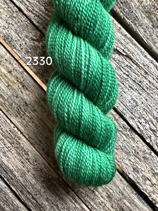 A skein of Koigu KPM & KPPPM Fingering Weight Yarn in vibrant green, labeled “2330,” rests on a weathered wooden surface with visible grain and gaps between the planks.