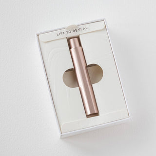 A refillable rose gold pen, The Wren, shaped like a sleek spray bottle, comes in a white box with a window and LIFT TO REVEAL on the flap. Its displayed on a plain white background.