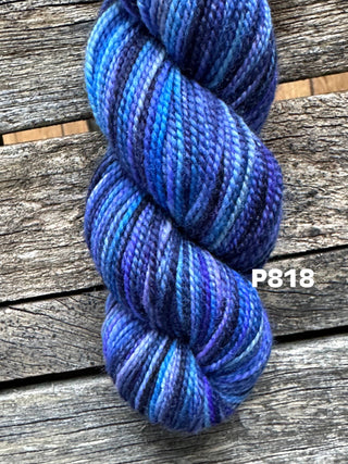 A skein from the KPM and KPPPM Variegated Color Collection in blue and purple lies on a weathered wooden surface. Tightly twisted for hand knitting, the yarn displays code P818 in white text beside it.