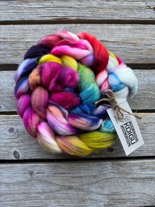 A Koigu Merino Roving Braid in pink, red, yellow, blue, green, and purple rests on a wooden surface with a branded tag attached—ideal for spinning fiber projects.