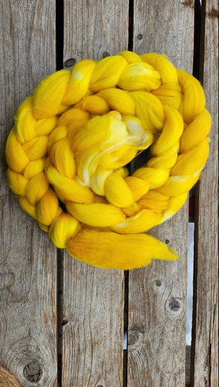 The Koigu Merino Roving Braid, a thick, bright yellow bundle with subtle color variations and white patches, is coiled in a circle on a weathered wooden surface.