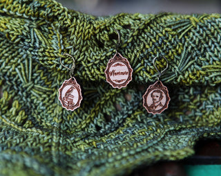 Three gothic vintage designs—a sailing ship, "Wettermete," and a portrait—are engraved on wooden Nevermore Knitting & Crochet Stitch Markers, shown hanging on green leaf-patterned knitted fabric.