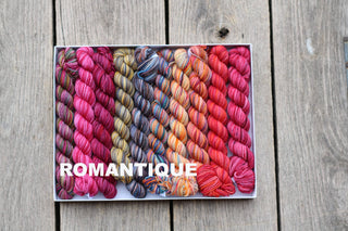 A white box labeled ROMANTIQUE on the side displays a curated Koigu Pencil Box yarn set with vibrant skeins in pink, purple, yellow, blue, and orange on a wooden surface.