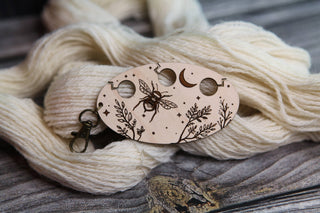 The Yarn Management Tool for Knitters is an oval wooden gauge engraved with a bee, moon, stars, and leaves. Resting on cream-colored yarn and set on a rustic wooden surface, this charming knitting tool features a convenient metal clip attached to the gauge.