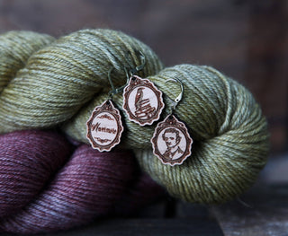 Two skeins of yarn (olive green and purple) plus three attached Nevermore Knitting & Crochet Stitch Markers with engraved designs: "Notemake," a hand holding knitting needles, and a person's portrait.