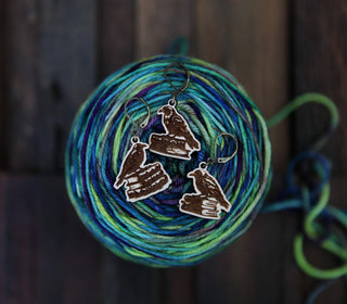 The Conspiracy of Ravens Knitting Progress Keepers feature three raven-shaped charms perched on stacked books, displayed atop blue-green variegated yarn on a dark wood surface—unique accessories for your next knitting project.