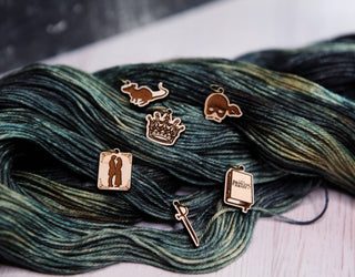 Five decorative charms—a crown, sword, book, kissing couple, and pirate hat—from the Princess Bride Knitting Stitch Markers set are arranged like unique knitting accessories on a skein of green and black yarn resting on a wooden surface. These charming stitch markers add both function and flair to your knitting projects.