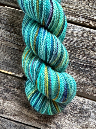 A KPM and KPPPM Variegated Color Collection Skein in teal, blue, yellow, and green rests on a weathered wooden surface—perfect for hand knitting projects.