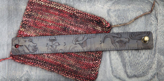 A triangular swatch of red and brown knit rests on grey, topped with the Mushroom Leather Ruler Cuff—a dark leather band etched with mushroom designs and numbered marks, ideal as a wearable ruler accessory.