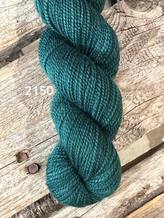 A skein of Koigu KPM & KPPPM Fingering Weight Yarn in deep teal (color 2150) lies on a weathered wooden surface, its tightly twisted merino strands ideal for knitting or crochet projects.