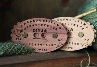 A close-up of two small cherry wood Ouija Board Row Counters, engraved with letters, numbers, YES, NO, and GOOD BYE, with green yarn in the background—perfect as a unique spirit board row tracker for knitters.