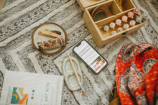 A flat lay of art supplies on a patterned rug, featuring the Walk in Nature Meditative Art Paint by Number Kit with paint pots, brushes, a phone displaying a wellness site, a ceramic dish with sage, and red patterned fabric for a calming ritual.