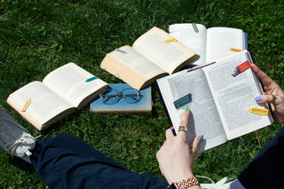 Open books and glasses on a grassy surface with a person reading one of the books.