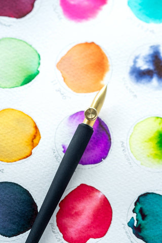 The One-Dip-Nib, a gold and black calligraphy dip pen, rests on watercolor paper beside vibrant circular paint swatches and handwritten color names.