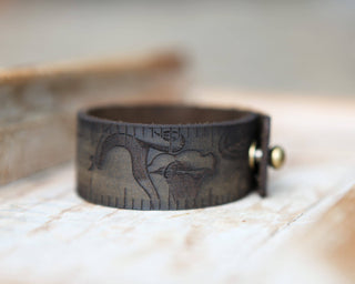 The Mushroom Leather Ruler Cuff is a wide, dark brown vegan leather bracelet featuring engraved ancient Egyptian-inspired designs and a metal button clasp, shown on a light wooden surface.