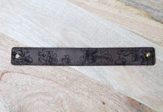 The Oak Leaves & Acorn Leather Ruler Cuff is a brown leather ruler with black markings, subtle leaf patterns, rounded corners, and metal studs—ideal as a wearable ruler cuff or shawl cuff, shown on a light wooden surface.