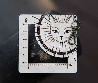 The Witchy Cat Gauge Swatch Ruler is a square wooden knitting ruler with inch and centimeter markings, an engraved cat face with sun rays, a metal clasp, and serves as a charming cat stitch marker for your crafts.