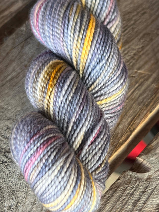 A close-up of a KPM and KPPPM Variegated Color Collection Skein in shades of grey, purple, yellow, and pink, neatly twisted on a rustic wooden surface—perfect for your next hand knitting project.