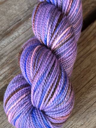 A skein from the KPM and KPPPM Variegated Color Collection, featuring tightly twisted merino strands in purple, blue, pink, and brown tones, rests on weathered wood—ideal for hand knitting with soft merino wool.