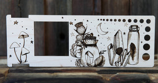 The Apothecary "Almost Everything" Ruler is a wooden yarn gauge tool with engraved mushrooms, an apothecary bottle with flowers, crystals, the moon, stars, a rectangular cutout on the left, and circular holes on the right.