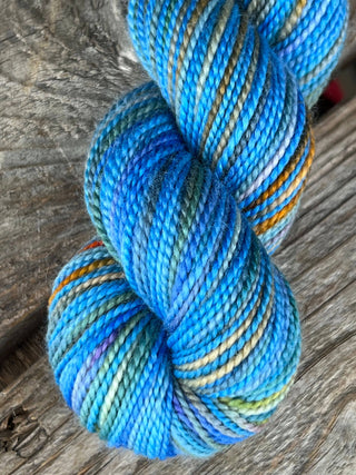 A close-up of a twisted KPM and KPPPM Variegated Color Collection Skein in blue, teal, green, and brown tones, perfect for hand knitting, displayed on a weathered wooden surface.