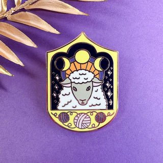 The Cosmic Sheep Enamel Pin is a limited edition accessory showcasing a cosmic sheep with a golden halo and yarn balls, set on a purple background adorned with moon phases and star motifs inspired by saint iconography.