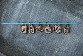 The Apothecary Bottle Stitch Marker Set features six small walnut wood markers shaped like jars with unique engravings, hanging from a blue knitting needle—a charming accessory for any knitting project.