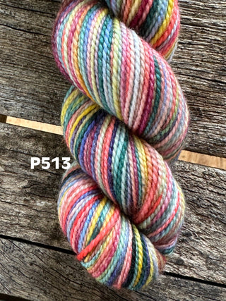 A skein from the KPM and KPPPM Variegated Color Collection, featuring multicolored stripes—red, blue, yellow, green, and pink (label P513)—rests on rustic wood. This soft merino wool is ideal for hand knitting projects.