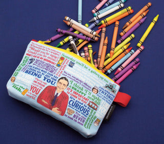 The Mister Rogers Zipper Pouch, featuring illustrated quotes and an image of Mister Rogers, rests on a dark blue surface with assorted crayons scattered around it.
