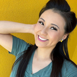 A woman with long dark hair smiles at the camera, wearing a green V-neck shirt and the Alien Joyride Earrings. She stands against a bright yellow textured wall with one hand behind her head.