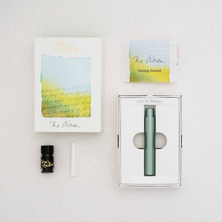 A flat lay of The Wren - Refillable Writing Pen starter kit showcases a decorated box, green pen with Japanese-made tip in a tray, small black bottle, white tube, and Getting Started card on a light surface.